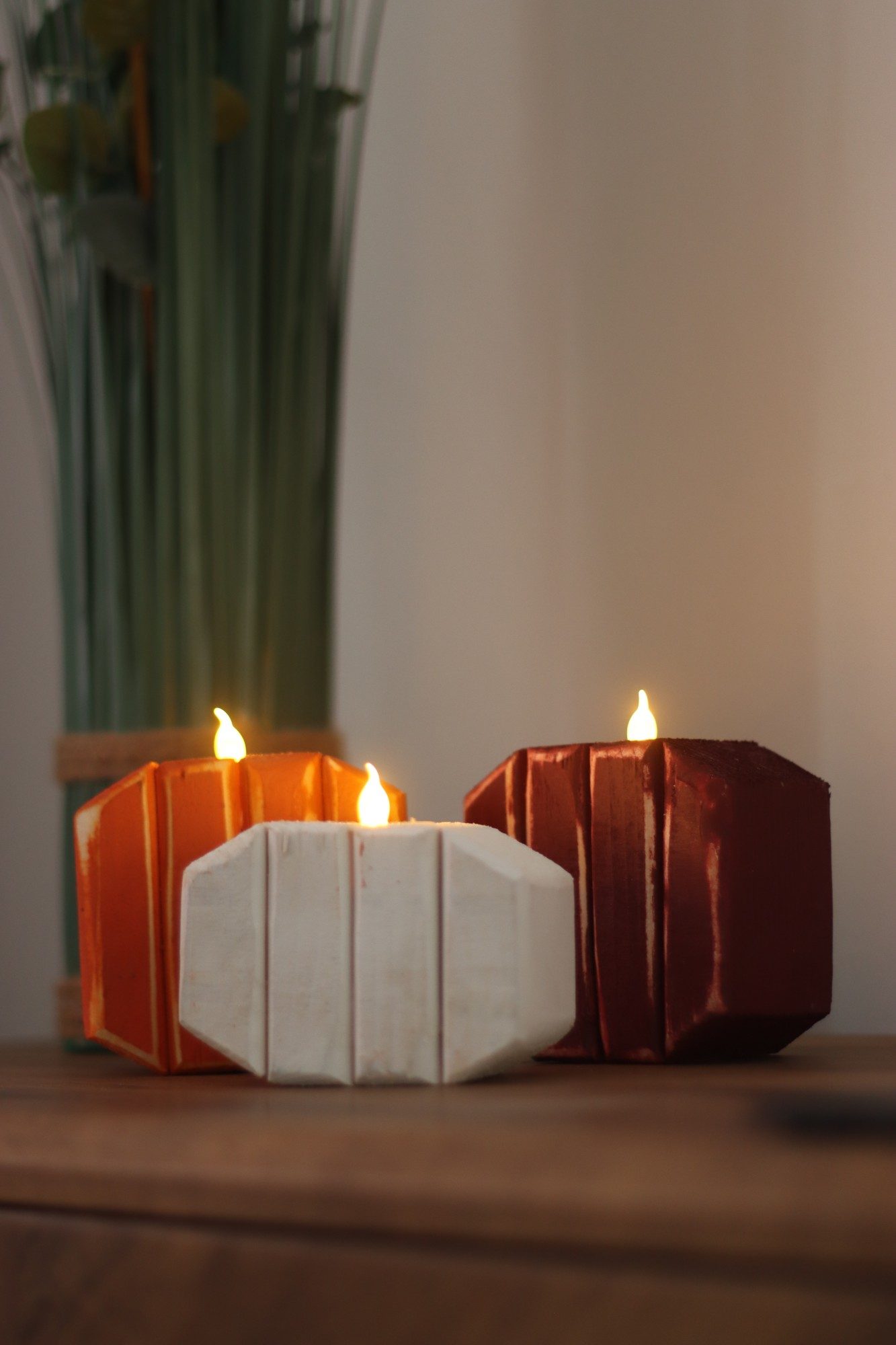 Three tea light pumpkins — orange, white and burgundy — glowing on a sideboard in the evening.